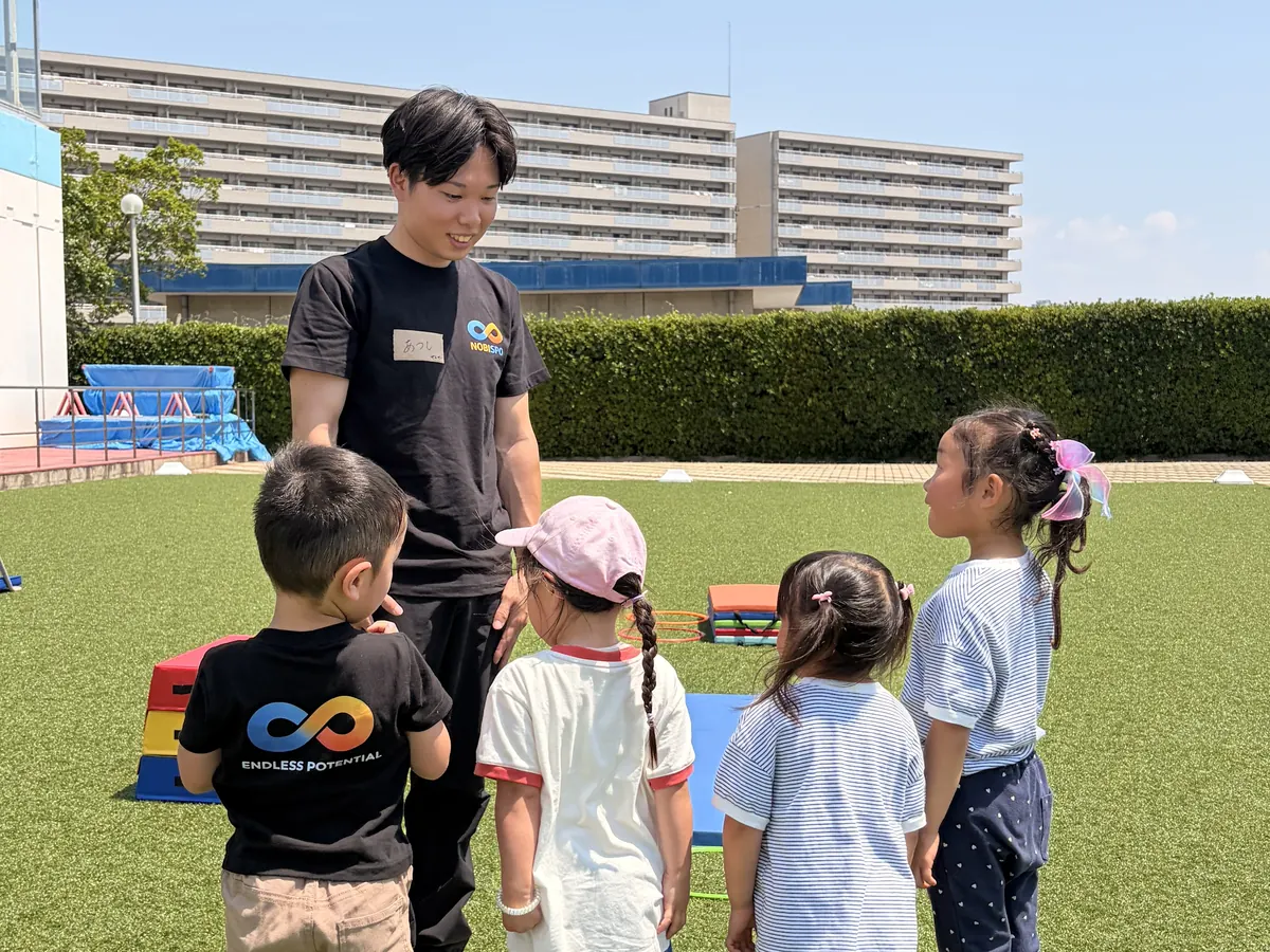 人工芝の運動フィールドでコーチが園児たちに笑顔で声をかける様子